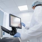 Pesquisadora de jaleco utiliza um monitor touchscreen em um laboratório moderno.