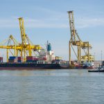 Navio cargueiro atracado em um terminal portuário, cercado por guindastes e contêineres.