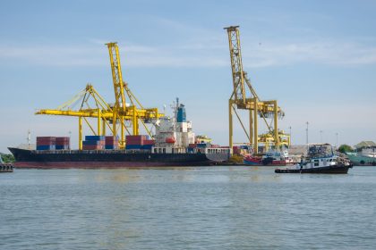 Navio cargueiro atracado em um terminal portuário, cercado por guindastes e contêineres.