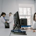 Pesquisadora trabalha em um computador em um laboratório, entre equipamentos e outros profissionais.