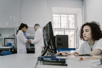 Pesquisadora trabalha em um computador em um laboratório, entre equipamentos e outros profissionais.