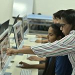 Professor e estudantes colaboram diante de computadores em um laboratório de informática.